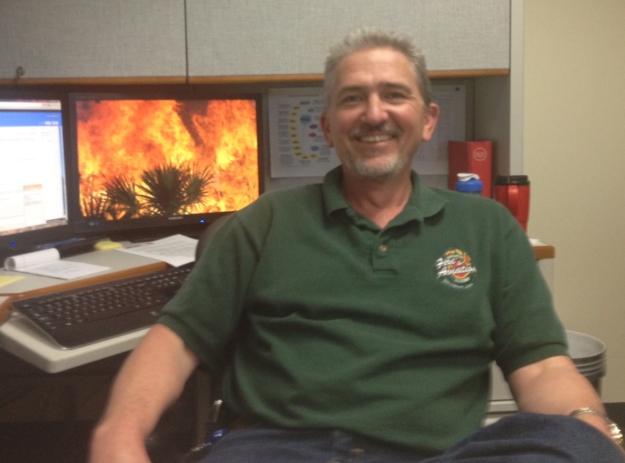 A man in a green shirt sits in front of a computer with flames in the background