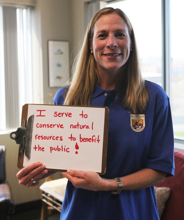 A woman with long reddish hair and a blue U.S. Fish and Wildlife Service emblem holds a sign saying " I serve to conserve natural resources to benefit the public!"